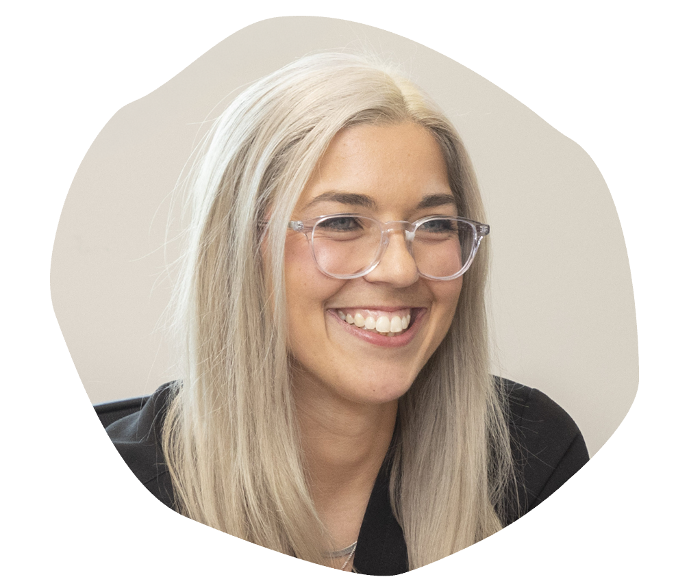 Smiling person with long blonde hair and glasses, wearing a dark top, against a neutral background. The mood is friendly and warm.