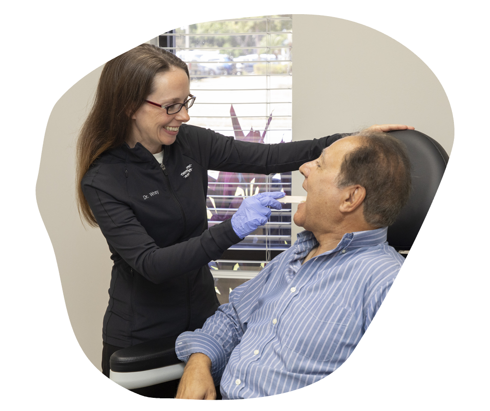 A dentist in glasses and gloves smiles while examining a male patient's mouth in a dental office. The room is bright with a view outside.