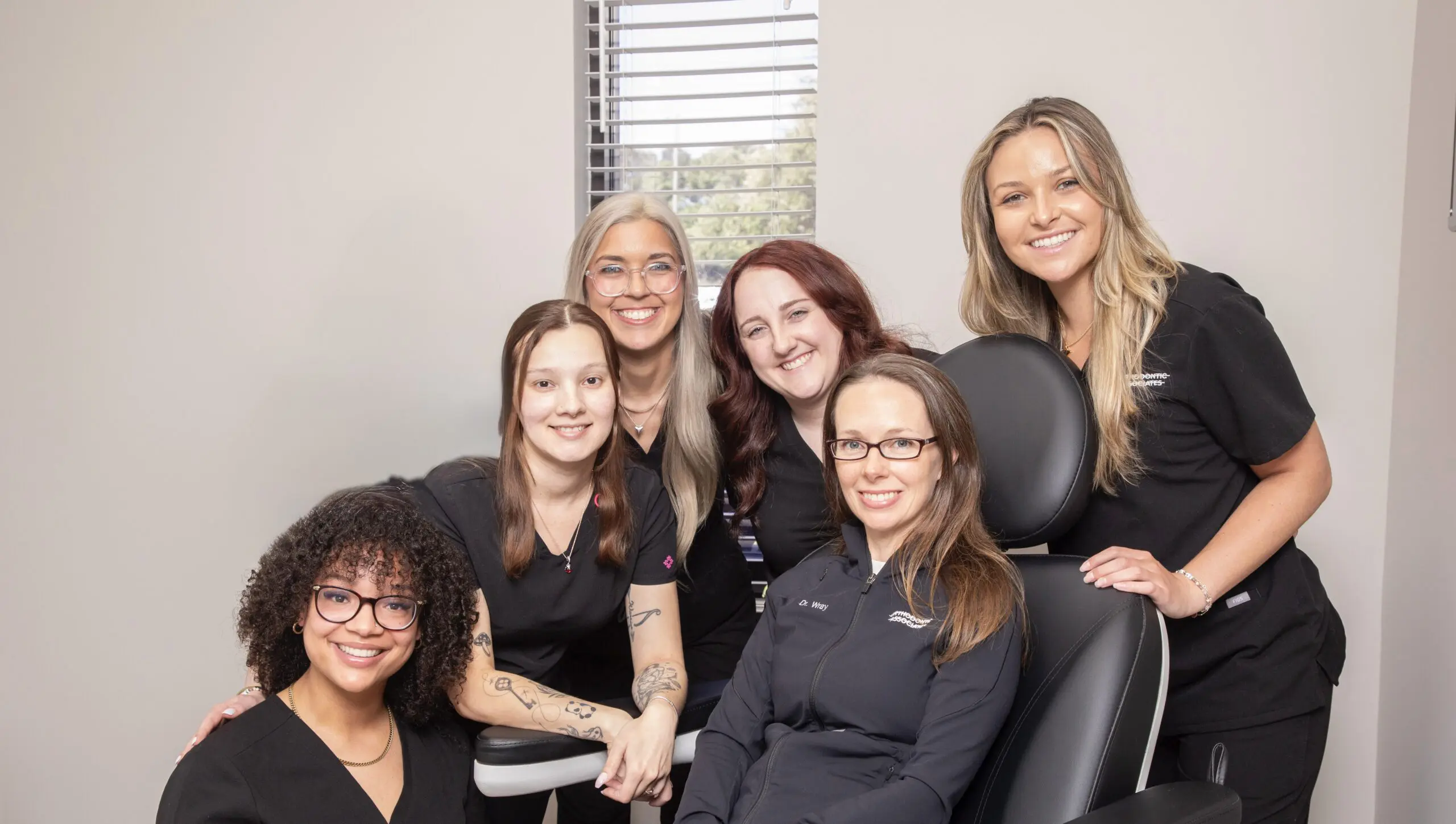 A group of six women in black attire, smiling warmly, pose together in an office setting. The atmosphere is friendly and professional.