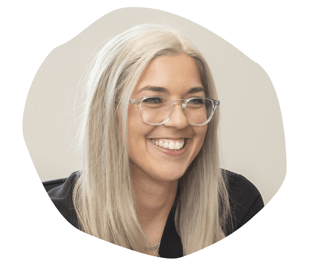 Smiling person with long blonde hair and glasses, wearing a dark top, against a neutral background. The mood is friendly and warm.