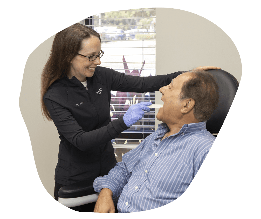 A dentist in glasses and gloves smiles while examining a male patient's mouth in a dental office. The room is bright with a view outside.