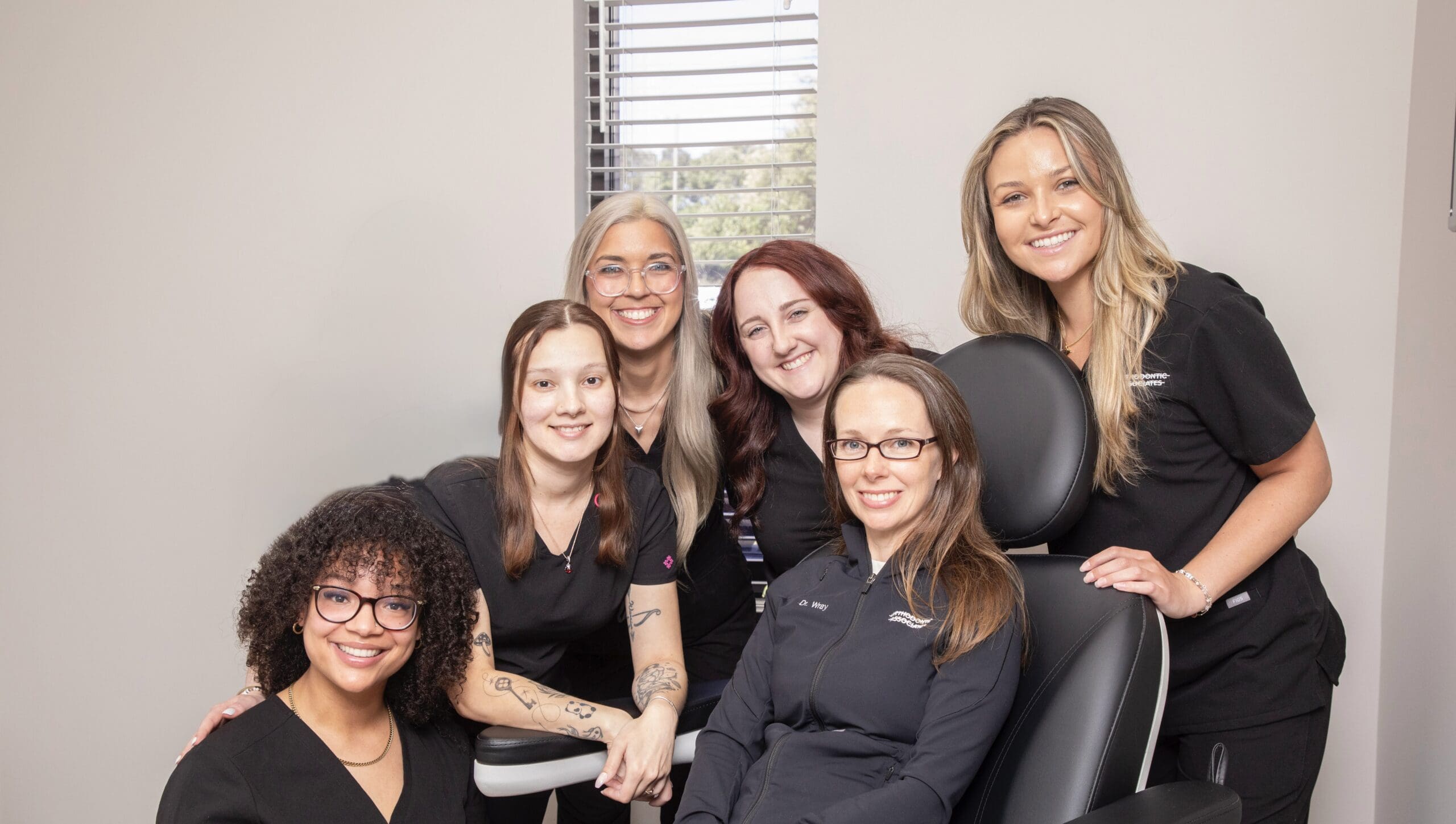 IMG_8183 A group of six women in black attire, smiling warmly, pose together in an office setting. The atmosphere is friendly and professional.